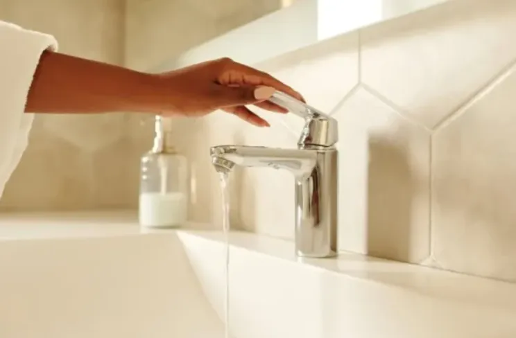 A person's hand turning a modern chrome faucet handle to run water in a bright, contemporary bathroom.