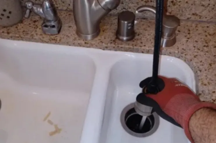 A gloved hand inserting a professional drain cleaning cable into a kitchen sink drain to clear a clog.