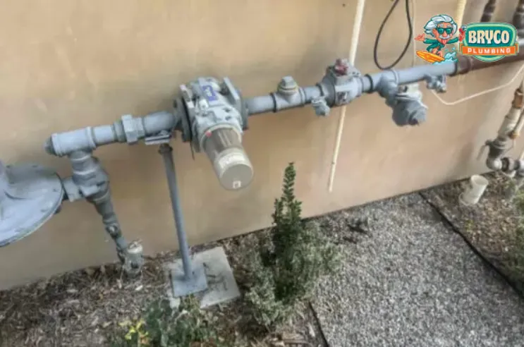 An outdoor installation of a gray natural gas meter and regulator assembly connected to pipes along a building's exterior wall.